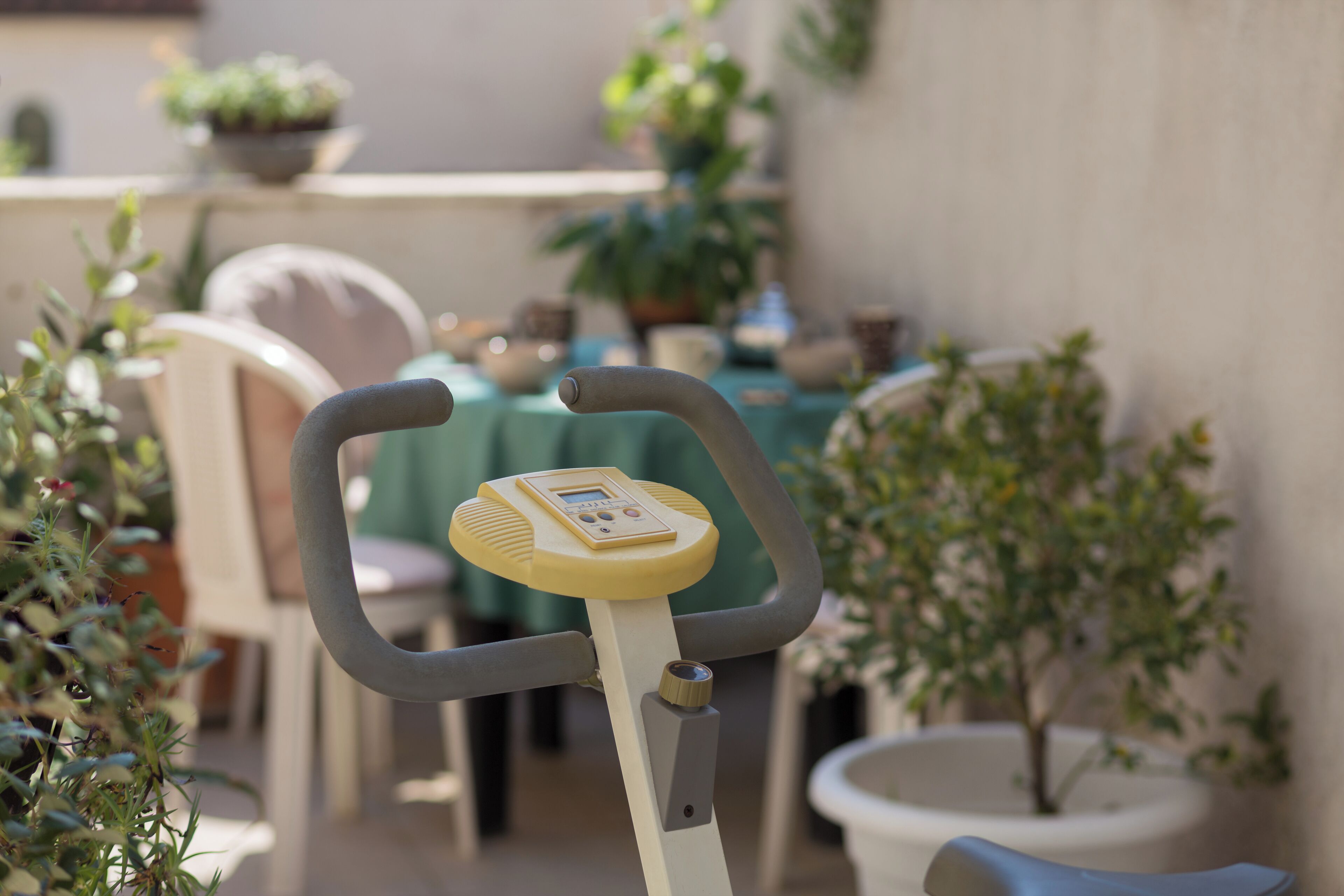 A stationary exercise bike with a digital display, positioned in an outdoor patio area with potted plants and a table with chairs.