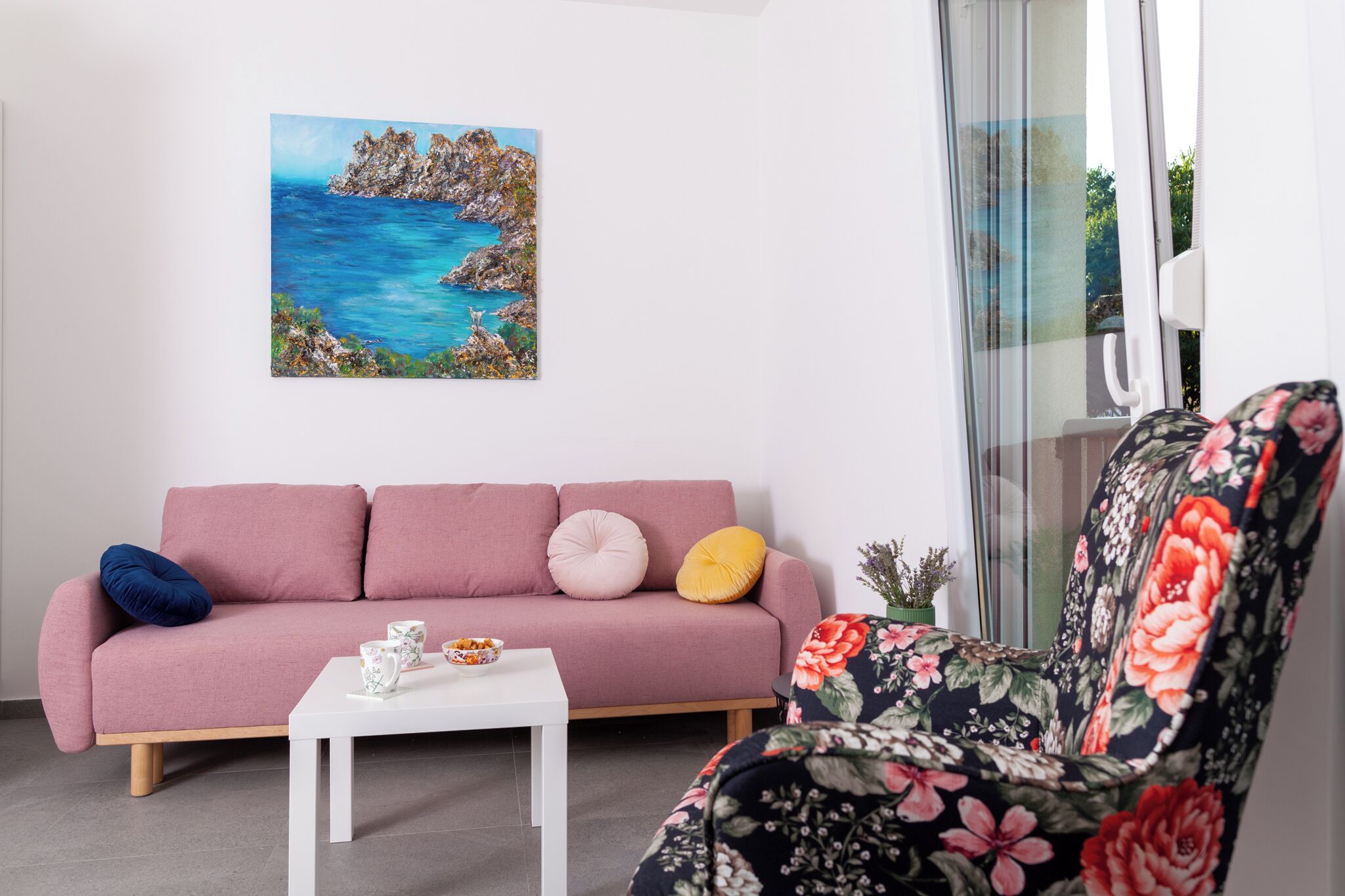Een woonkamer met een roze bank, een witte salontafel en een fauteuil met bloemenprint. Op de muur hangt een schilderij van een kustlandschap.