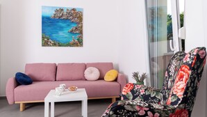 Una sala de estar con un sofá rosa, una mesa de centro blanca y un sillón estampado con motivos florales. Un cuadro de una escena costera adorna la pared.