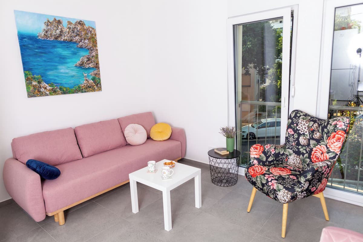 Un canapé rose avec des coussins, une table basse blanche avec deux tasses, un fauteuil à motifs floraux et un tableau représentant un paysage côtier.