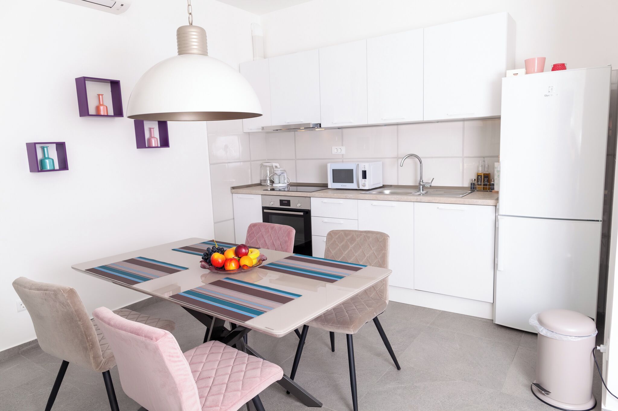 Een moderne keuken met witte kastjes, een magnetron en een spoelbak. Er staat een eettafel met stoelen en een schaal met fruit erop.