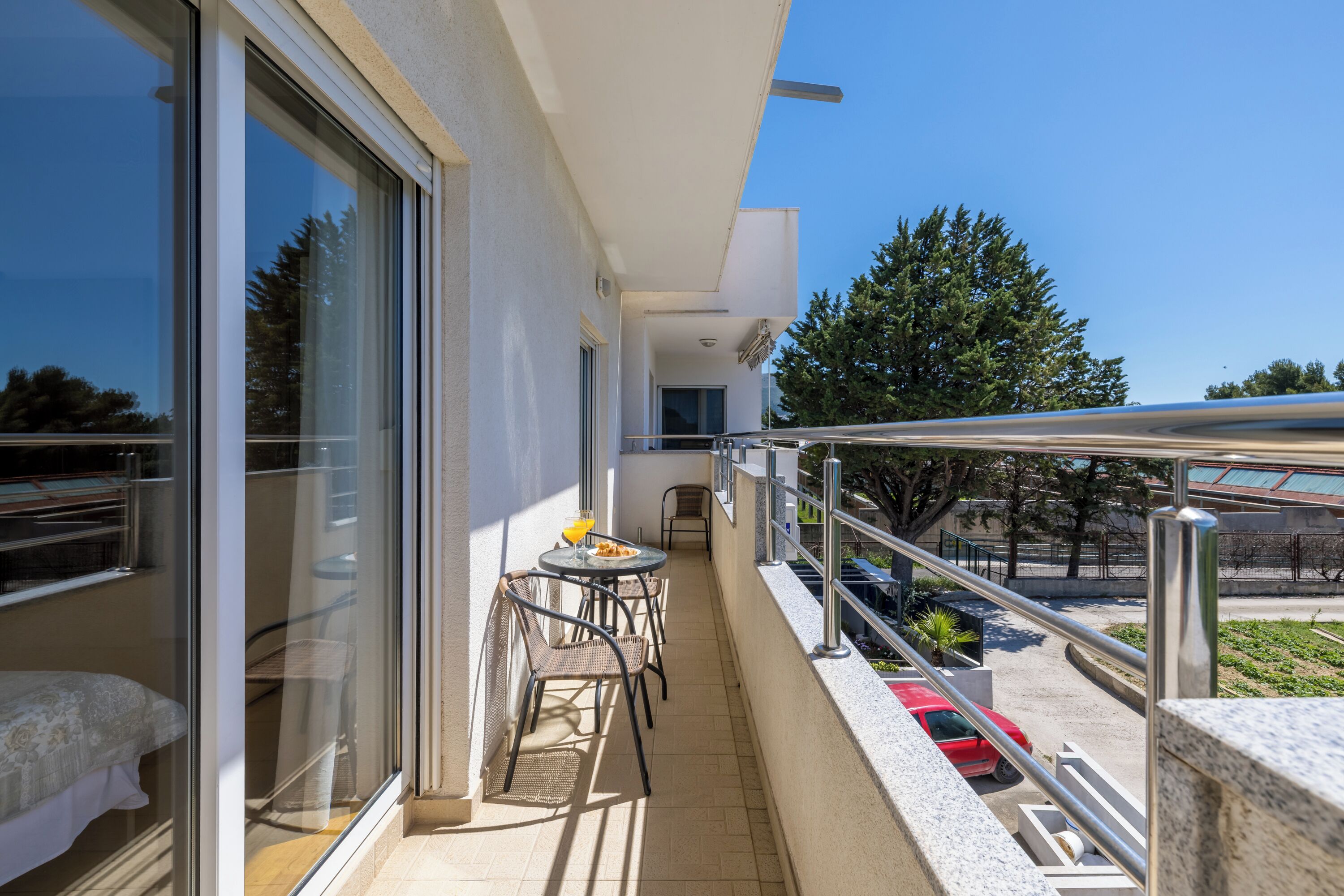 A balcony with a glass door, a small table with chairs, and a red car parked outside.