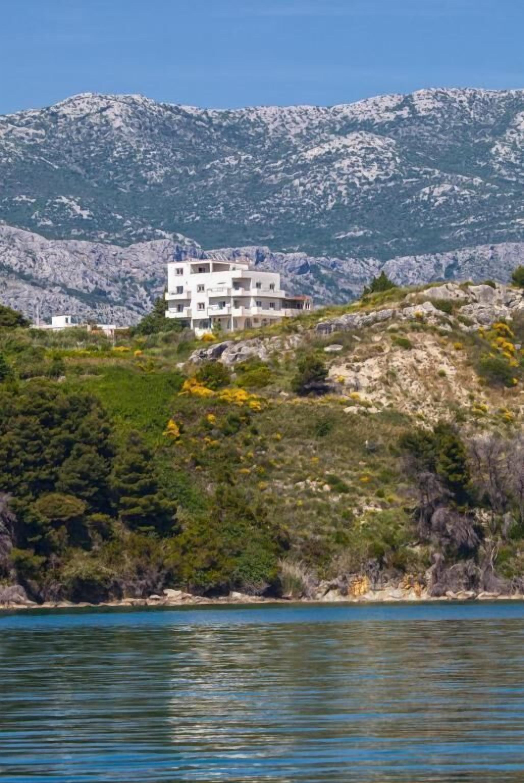 Ein Gebäude an der Küste mit mehreren Stockwerken, gelegen auf einer Anhöhe mit Blick auf das Meer und Berge im Hintergrund.