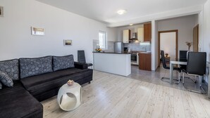 A modern living room with a dark sofa, a white coffee table, and a small pet bed. A kitchen with wooden cabinets and a dining area with a white table and black chairs is visible in the background. - Casa Di Tenor (Stobreč)