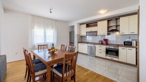A modern kitchen with white cabinets, stainless steel appliances, and a dining area with wooden furniture and a fruit bowl. - Casa Di Tenor (Stobreč)