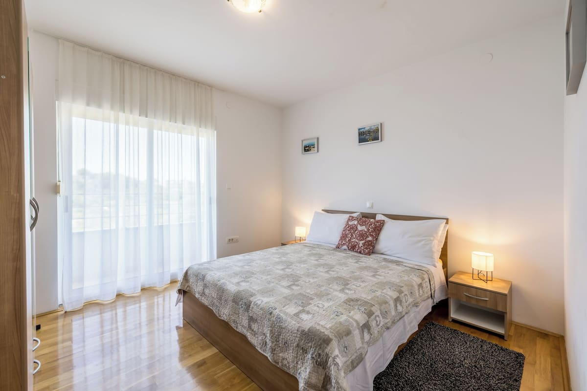 Ein Schlafzimmer mit einem großen Bett, einem Nachttisch mit Lampe, einem Fenster mit durchsichtigen Vorhängen und einem kleinen Bild an der Wand.