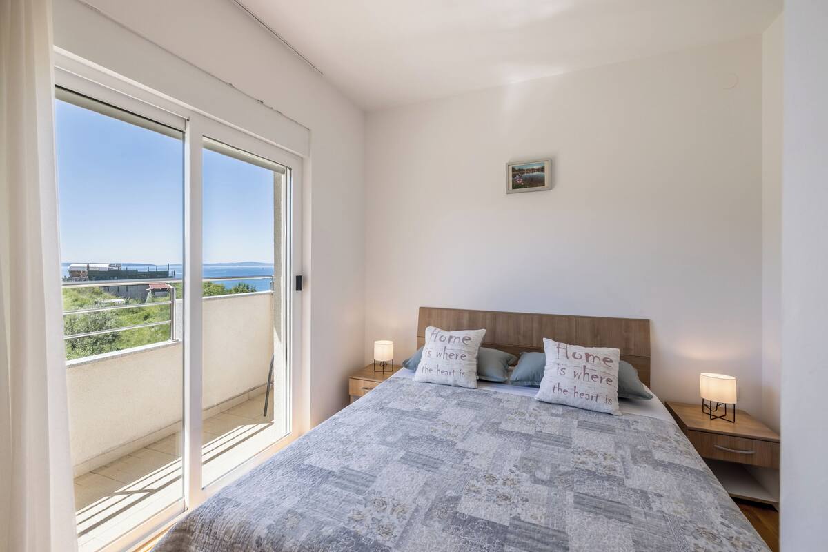 Ein Schlafzimmer mit einem großen Bett, zwei Nachttischen mit Lampen, einem kleinen Bild an der Wand und einer Glastür, die auf einen Balkon mit Blick auf das Meer und das entfernte Land führt.