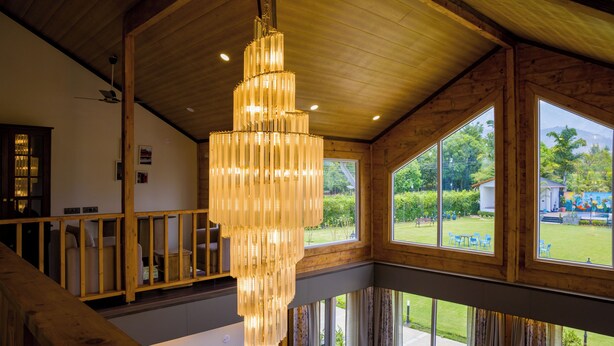 A spacious room with a large chandelier, wooden beams, and large windows offering a view of the outdoors. - ELIVAAS Esh Birch  (Palampur)