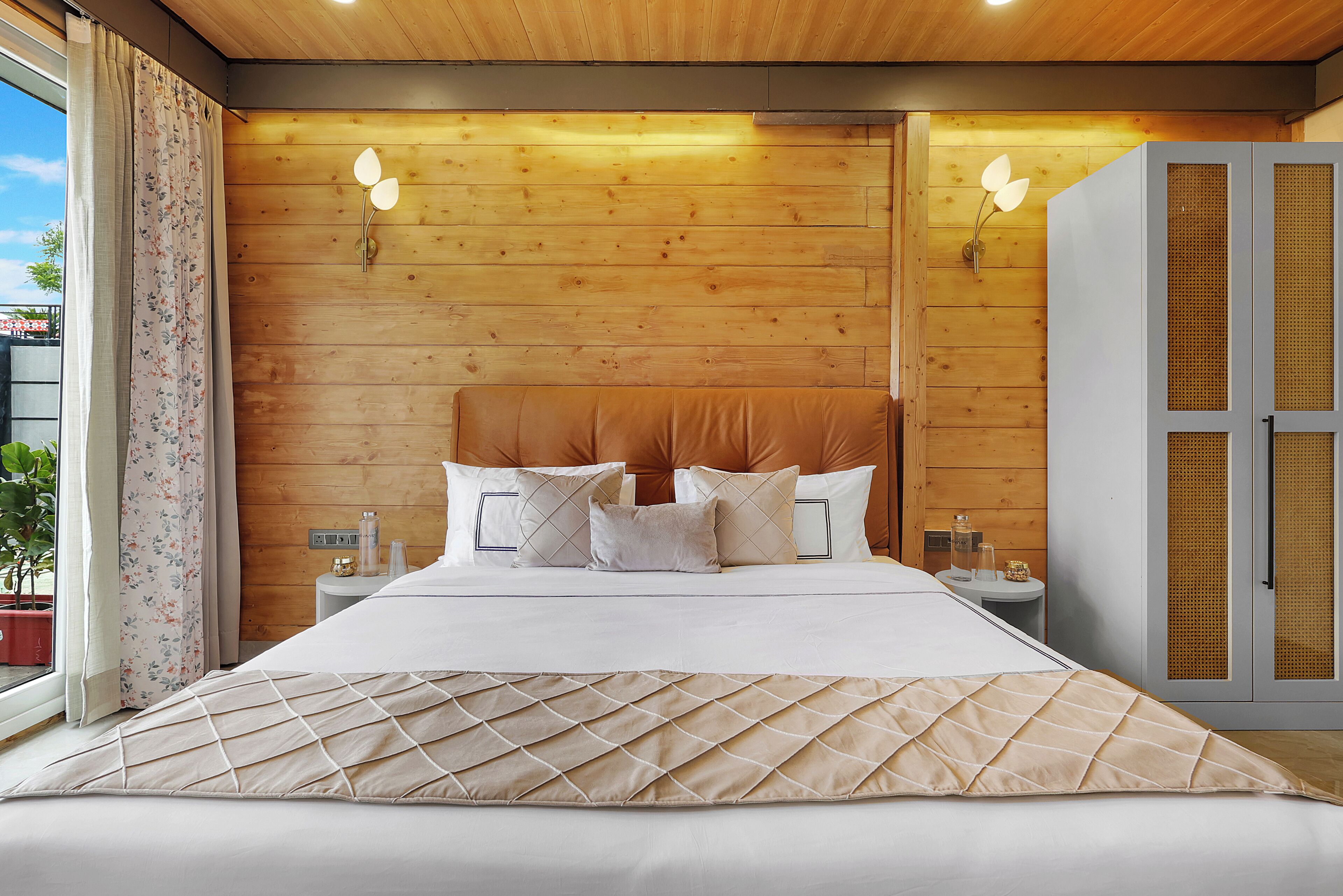 Un dormitorio moderno con cabecera de madera, una cama grande con cubrecama acolchado, dos mesitas de noche con lámparas y vistas al exterior a través de una ventana con cortinas.