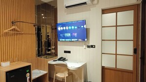 A hotel room with a wooden desk, a chair, a small fridge, a TV mounted on the wall, and a sliding door. - SK Village (Bangkok)