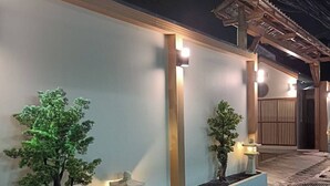 A well-lit exterior of a building with a modern design, featuring a low wall, two small trees, and a traditional Japanese lantern. - SK Village (Bangkok)