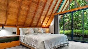Un dormitorio con techo de madera, una cama grande con ropa de cama estampada, una mesita de noche y vistas a la vegetación a través de amplias ventanas.