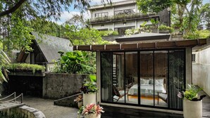 Un espacio moderno para sentarse al aire libre con cerramiento de vidrio, rodeado de abundante vegetación y una terraza de madera.