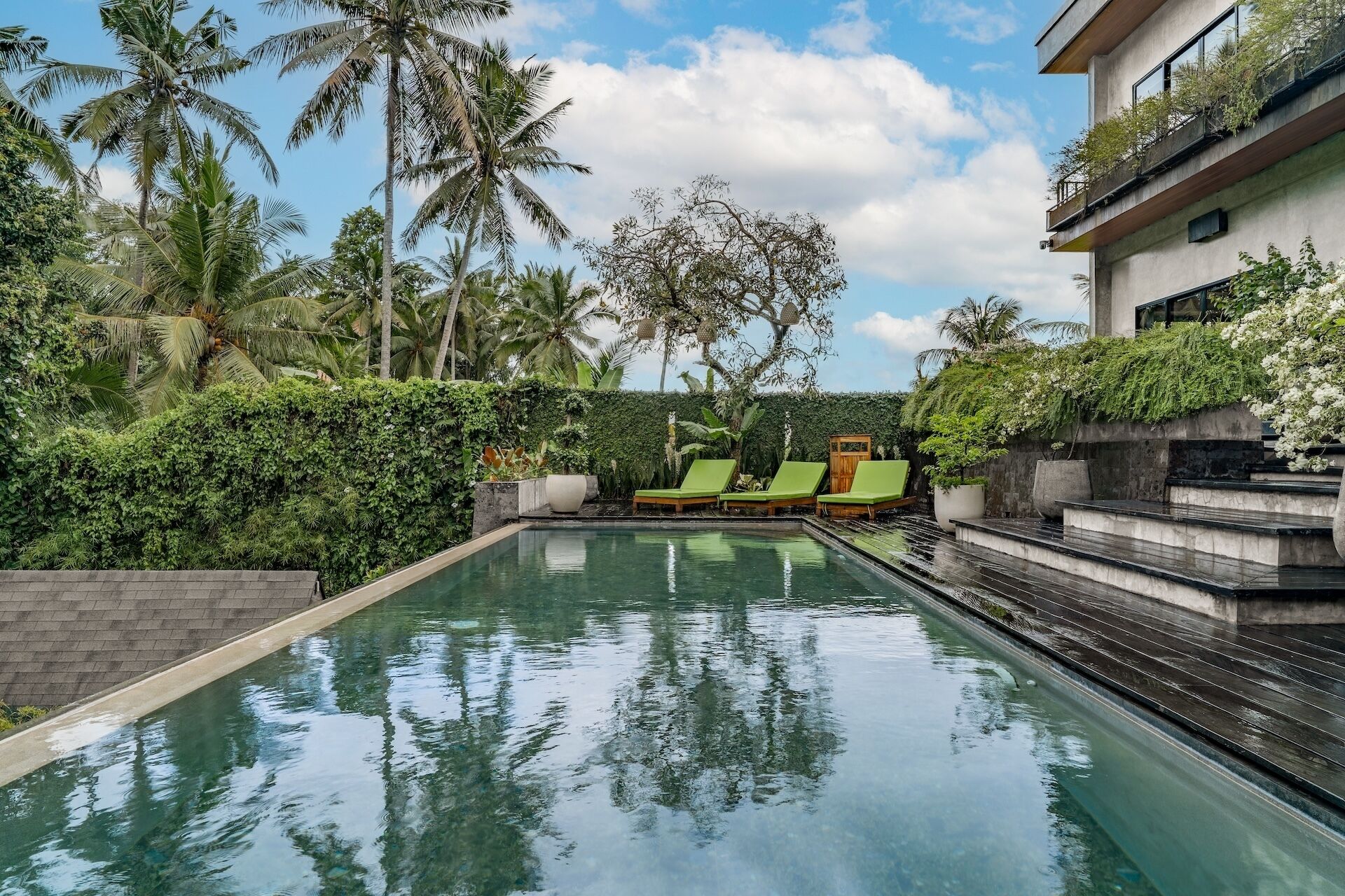 A swimming pool with lounge chairs, surrounded by tropical vegetation and a modern building.