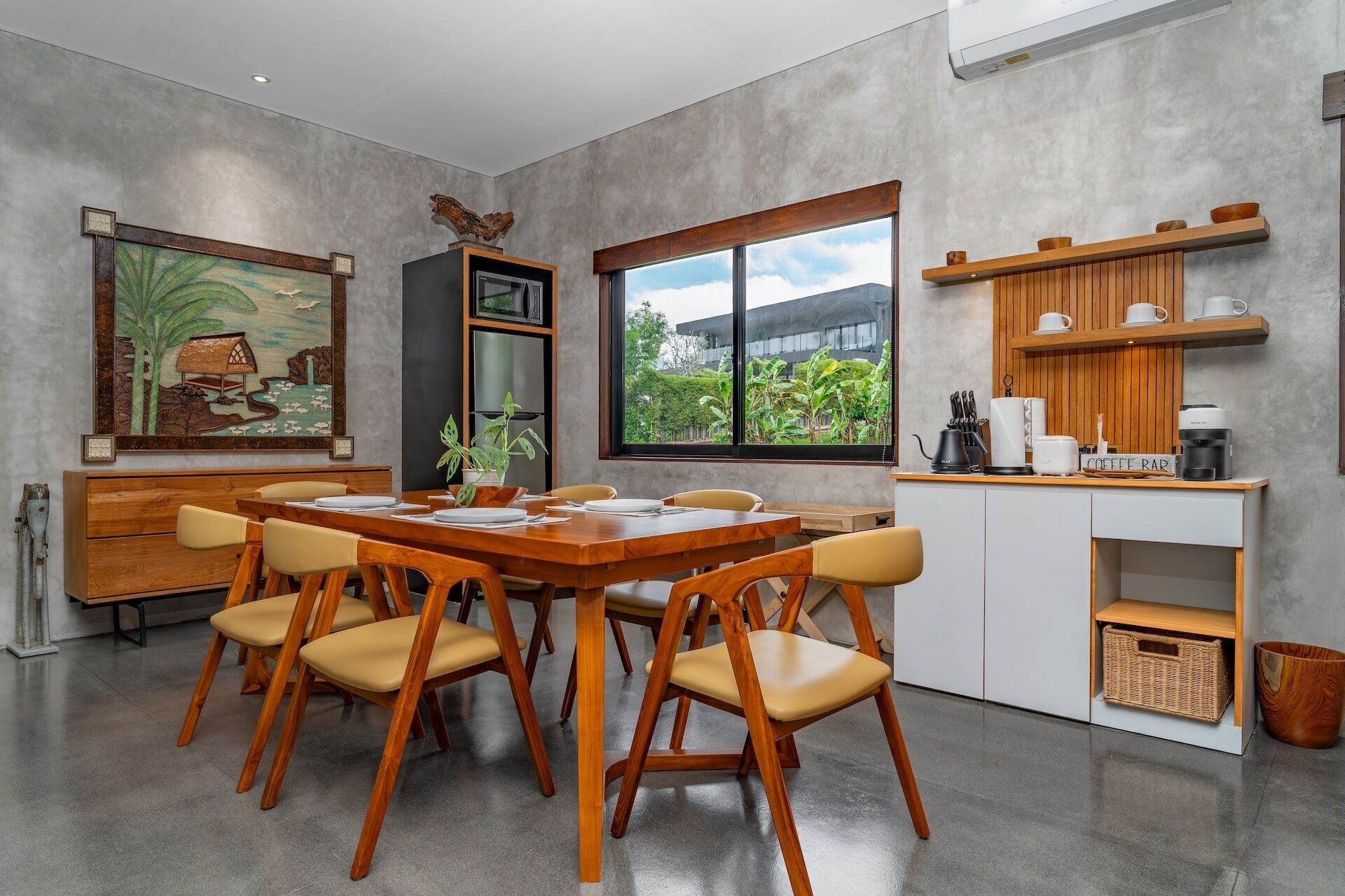 Een moderne eethoek met een houten tafel en stoelen, een koelkast, een koffiehoek en uitzicht op groen buiten.