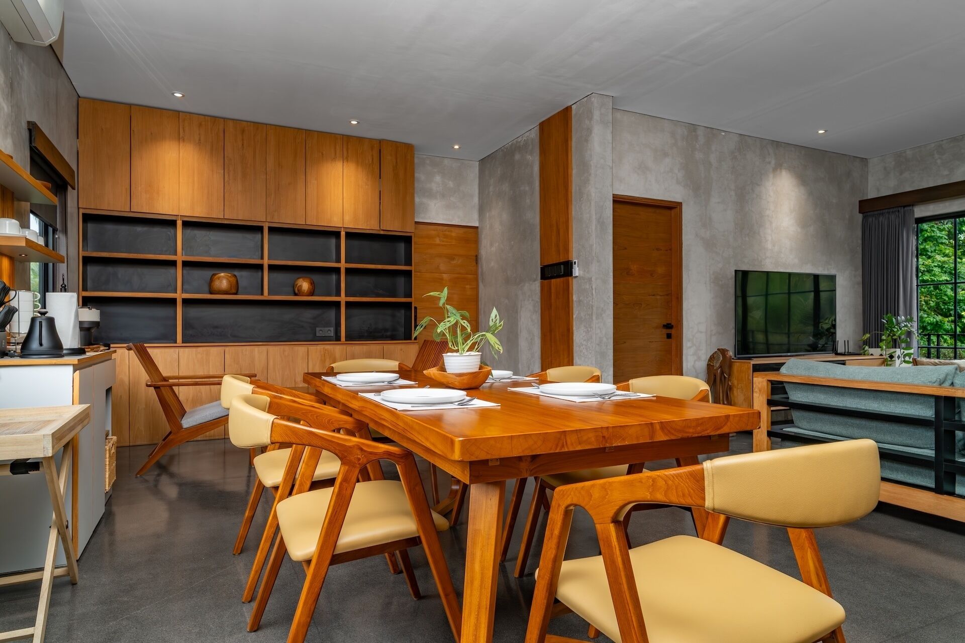 A modern dining area with a wooden table and chairs, a built-in shelf unit, and a television in the background.