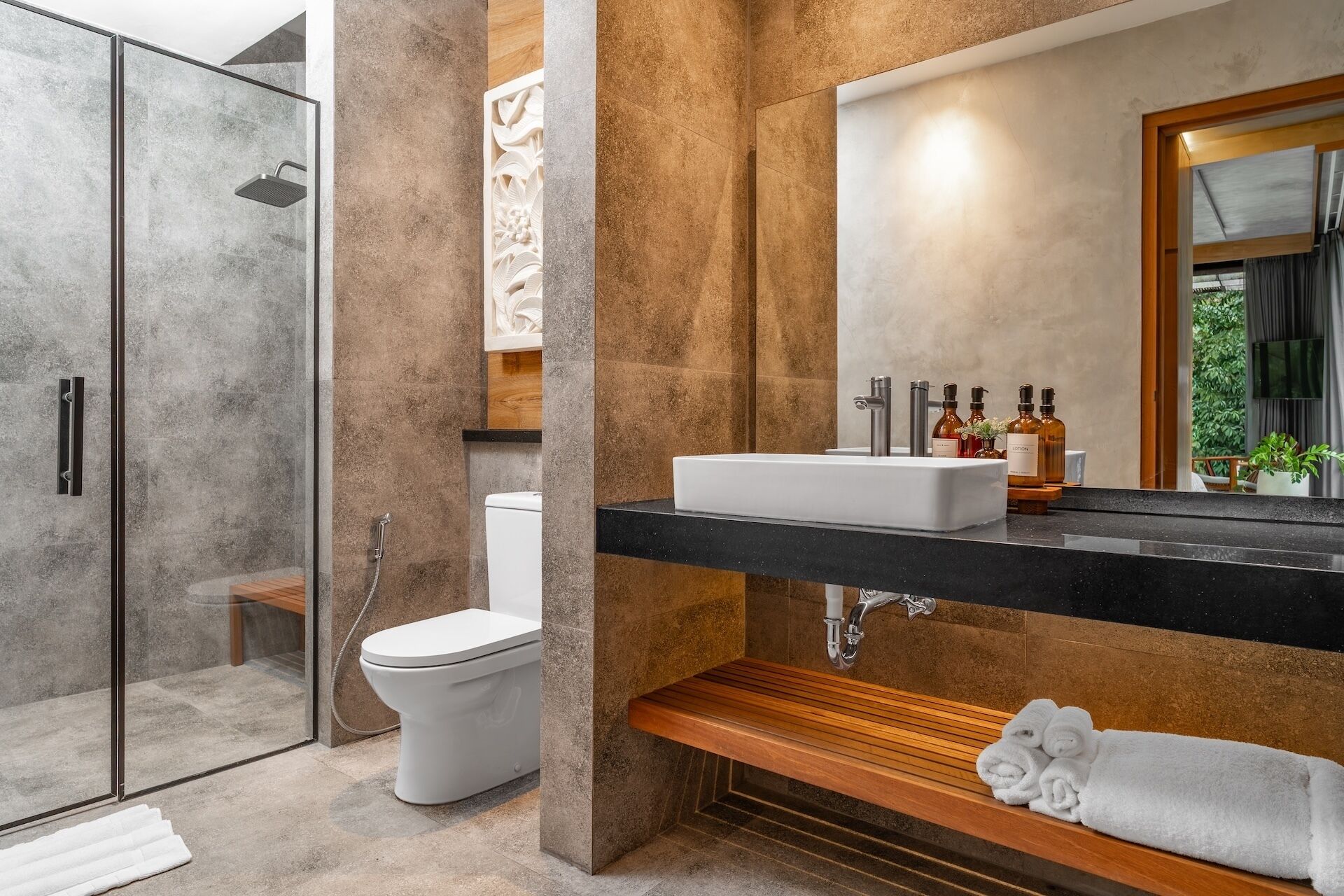 Une salle de bain moderne avec une douche à parois de verre, des toilettes blanches, un banc en bois et un lavabo avec des articles de toilette.