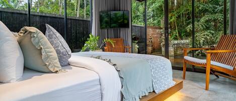 A modern bedroom with a large bed, a TV, and a view of lush greenery.