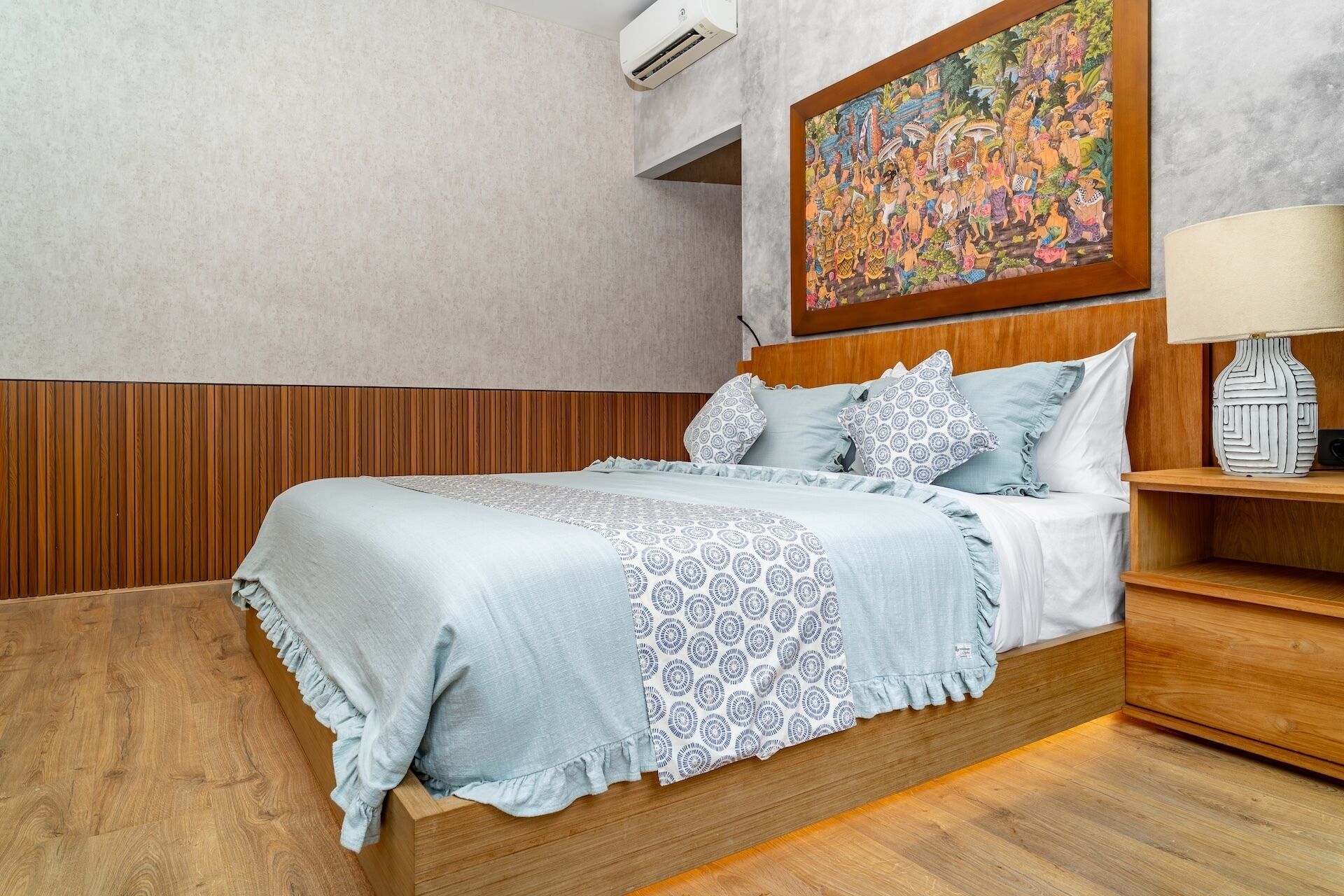 Una cama bien hecha con una colcha estampada y almohadas, un cabecero de madera, un aire acondicionado montado en la pared y un cuadro colorido sobre la cama.