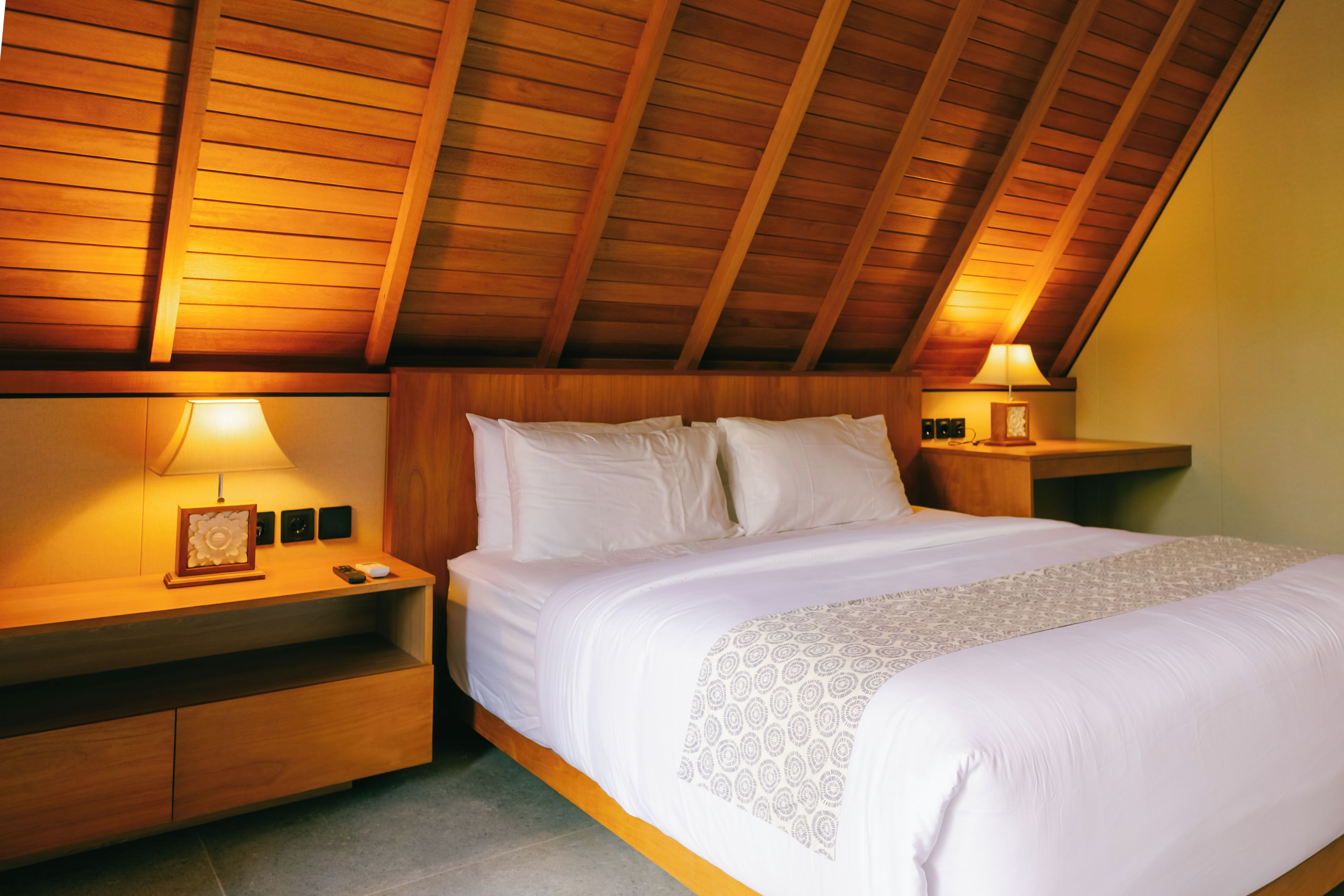 A bedroom with a sloped ceiling, wooden headboard, a bed with white linens, two bedside tables with lamps, and a nightstand with a picture frame.