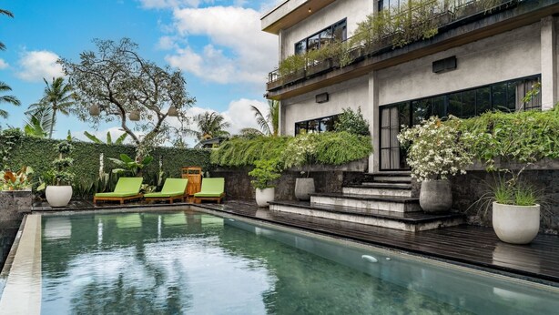 Un área de piscina moderna con agua cristalina, sillas de descanso verdes y un edificio con balcones y plantas.