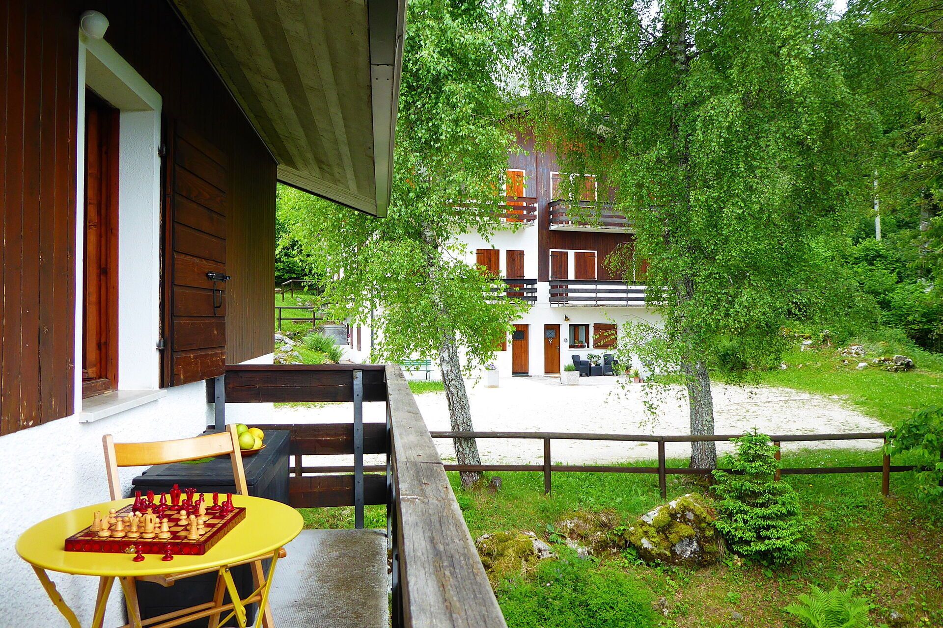 Une terrasse en bois avec un jeu d’échecs et une chaise, donnant sur un bâtiment blanc doté d’un balcon, le tout entouré de verdure.