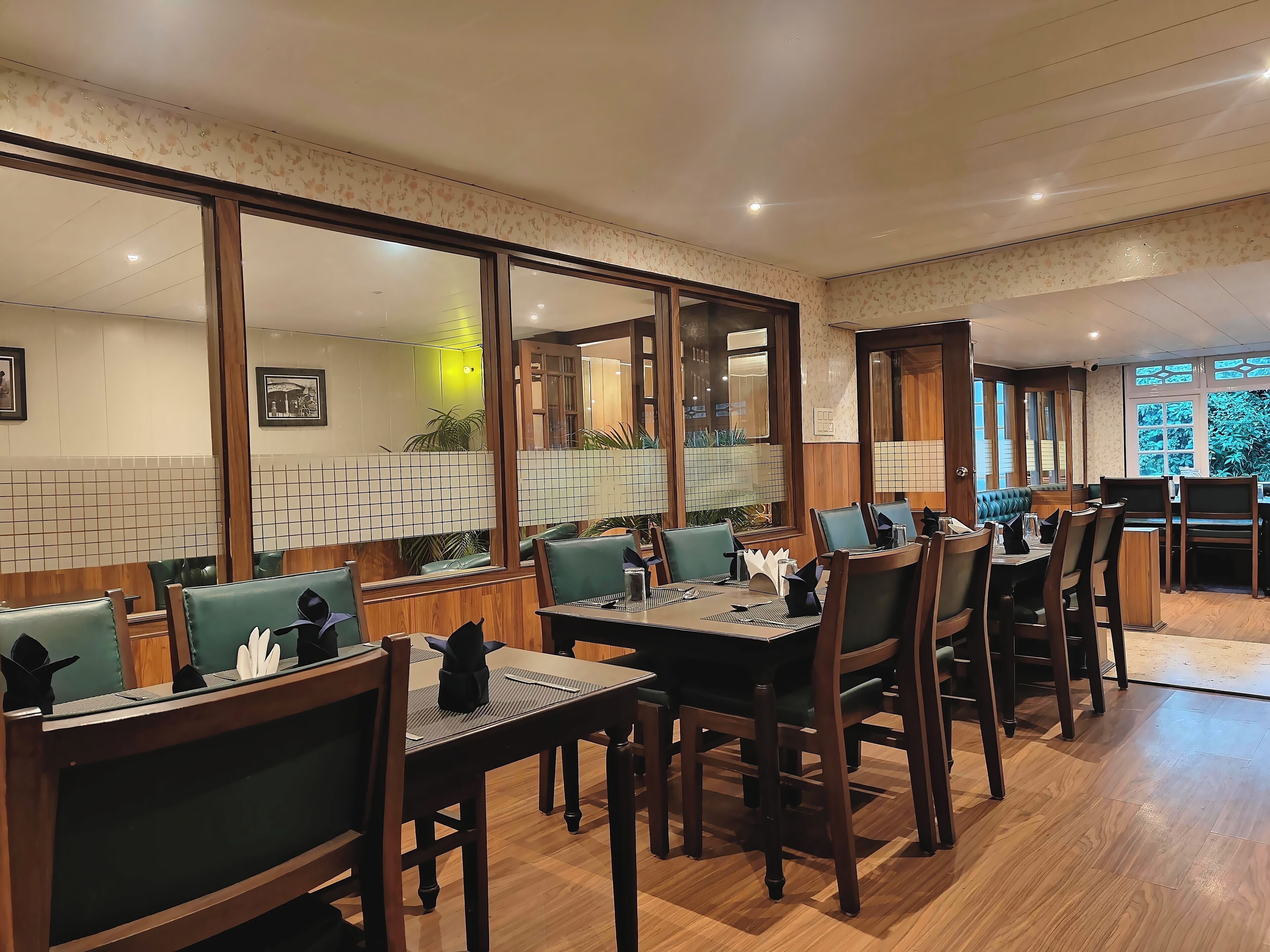 A restaurant with wooden tables and chairs, green cushioned booths, and large windows with a view of greenery.