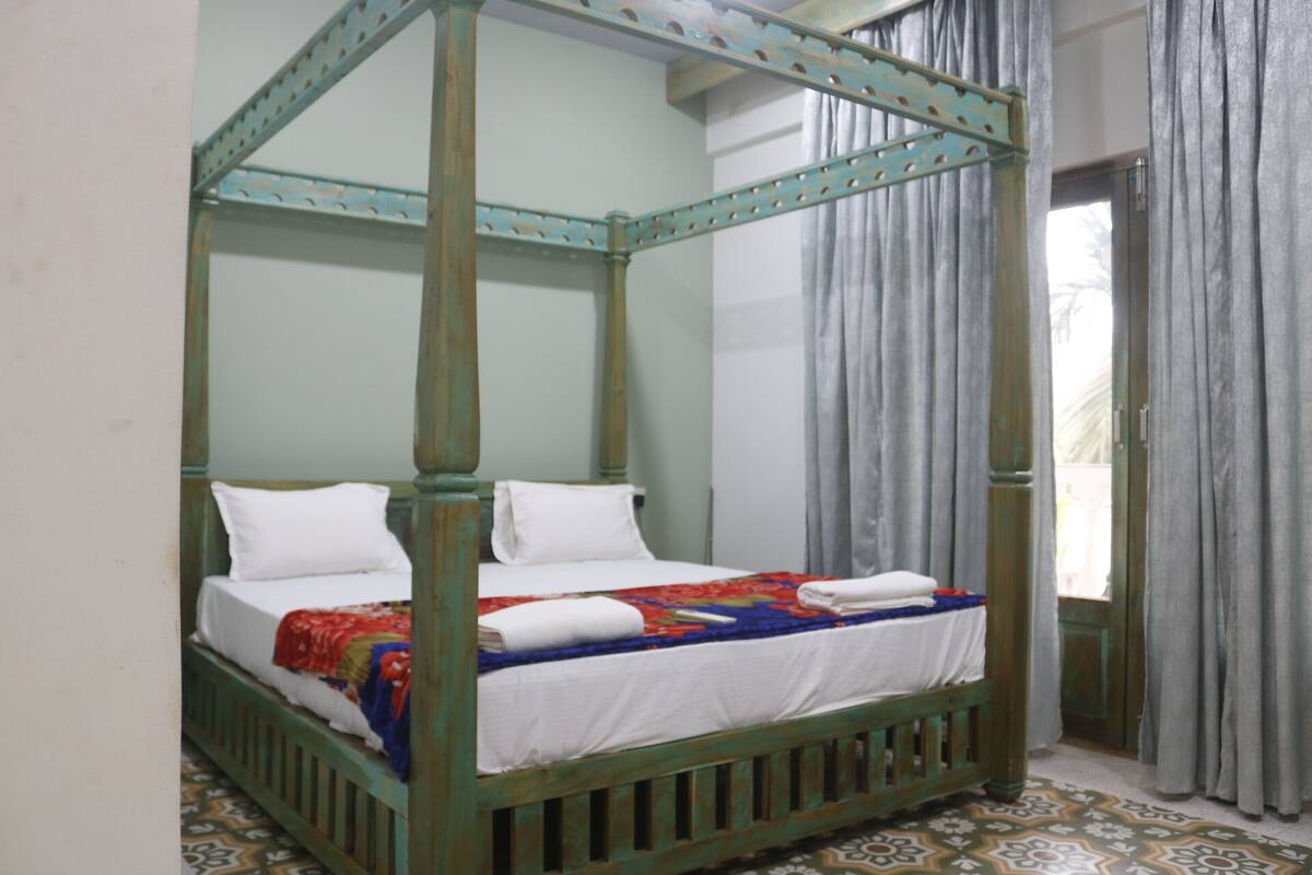 Un lit à baldaquin avec du linge de lit blanc et une couverture colorée, un plancher en bois orné d’un motif géométrique et une fenêtre avec des rideaux gris.