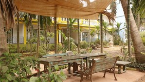 Un área de estar al aire libre con una mesa de madera y bancos, rodeada de vegetación y un edificio amarillo al fondo.