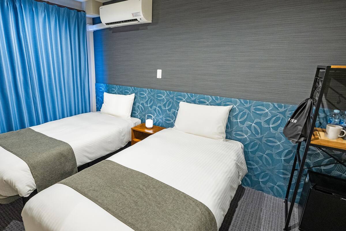 A hotel room with two beds, a nightstand with a lamp, a wall-mounted air conditioner, and a blue curtain.