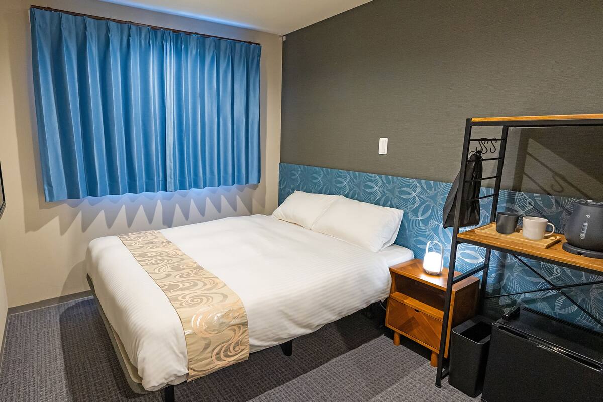 A hotel room with a bed, a bedside table with a kettle, a tray with a mug and a cup, and a blue curtain.