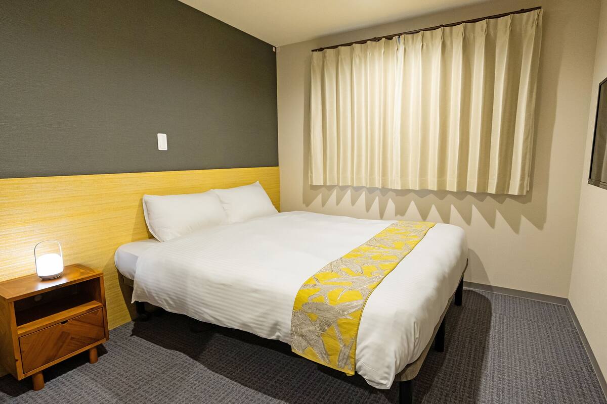 A hotel room with a bed, a bedside table with a lamp, a wooden nightstand, and a large window with curtains.
