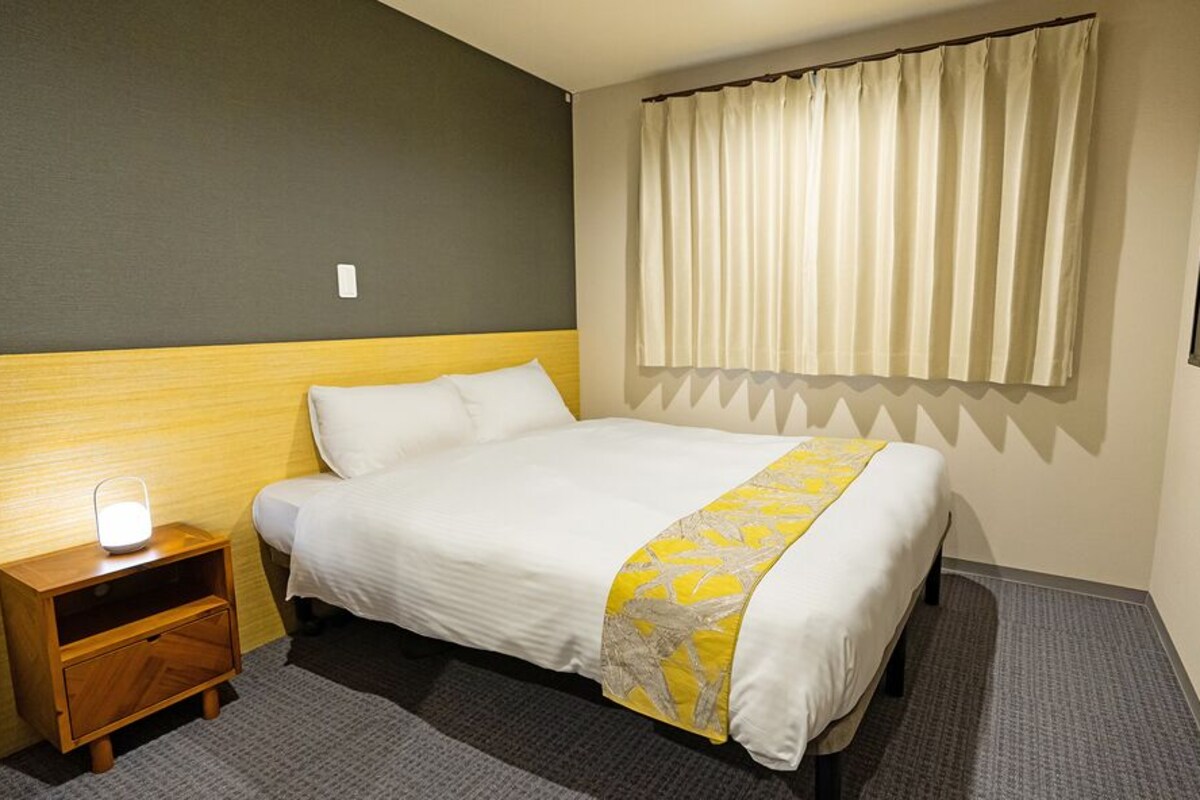 A hotel room with a bed, a nightstand, a lamp, a window with curtains, and a wall-mounted TV.