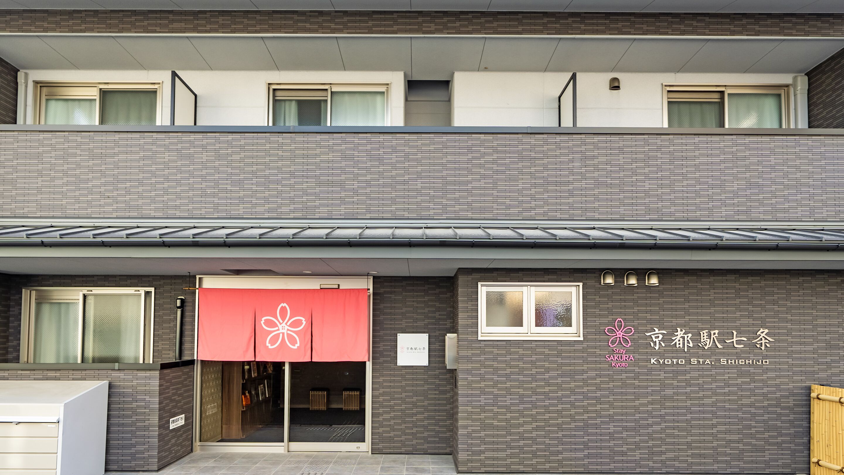 A hotel with a modern facade, featuring a red awning with a floral logo, and signage indicating it is located in Kyoto.