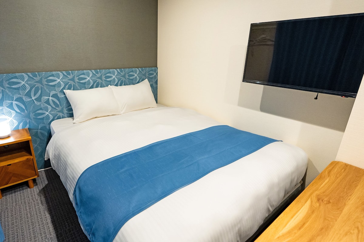 A neatly made bed with a blue and white bedspread, a wooden nightstand with a lamp, and a blue curtain.