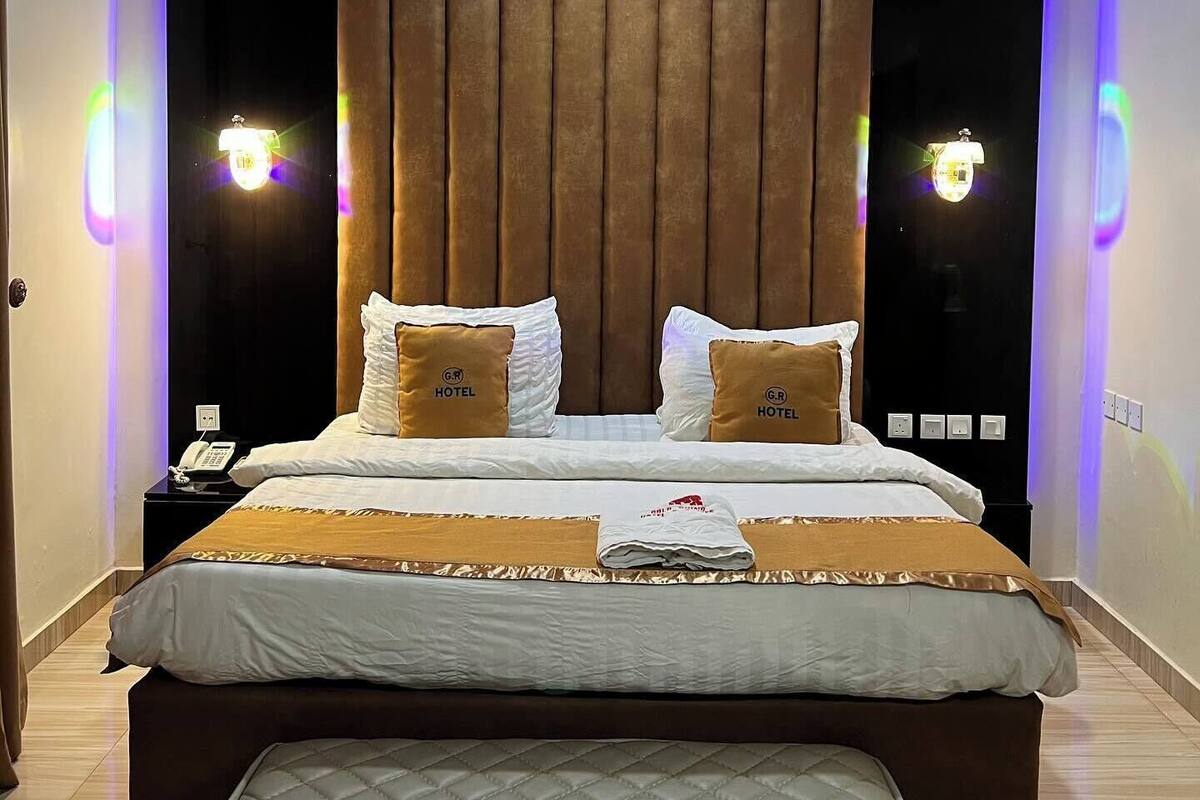 Habitación de hotel moderna con una cama grande, dos almohadas con el logotipo del hotel, una mesita de noche con un teléfono y un banco al pie de la cama.