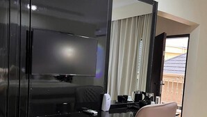 A modern bedroom with a black vanity, a large mirror, a flat-screen TV, a white bedside table, a pink chair, and a view of a wooden deck outside. - Gold Rhino Hotel (Enugu)