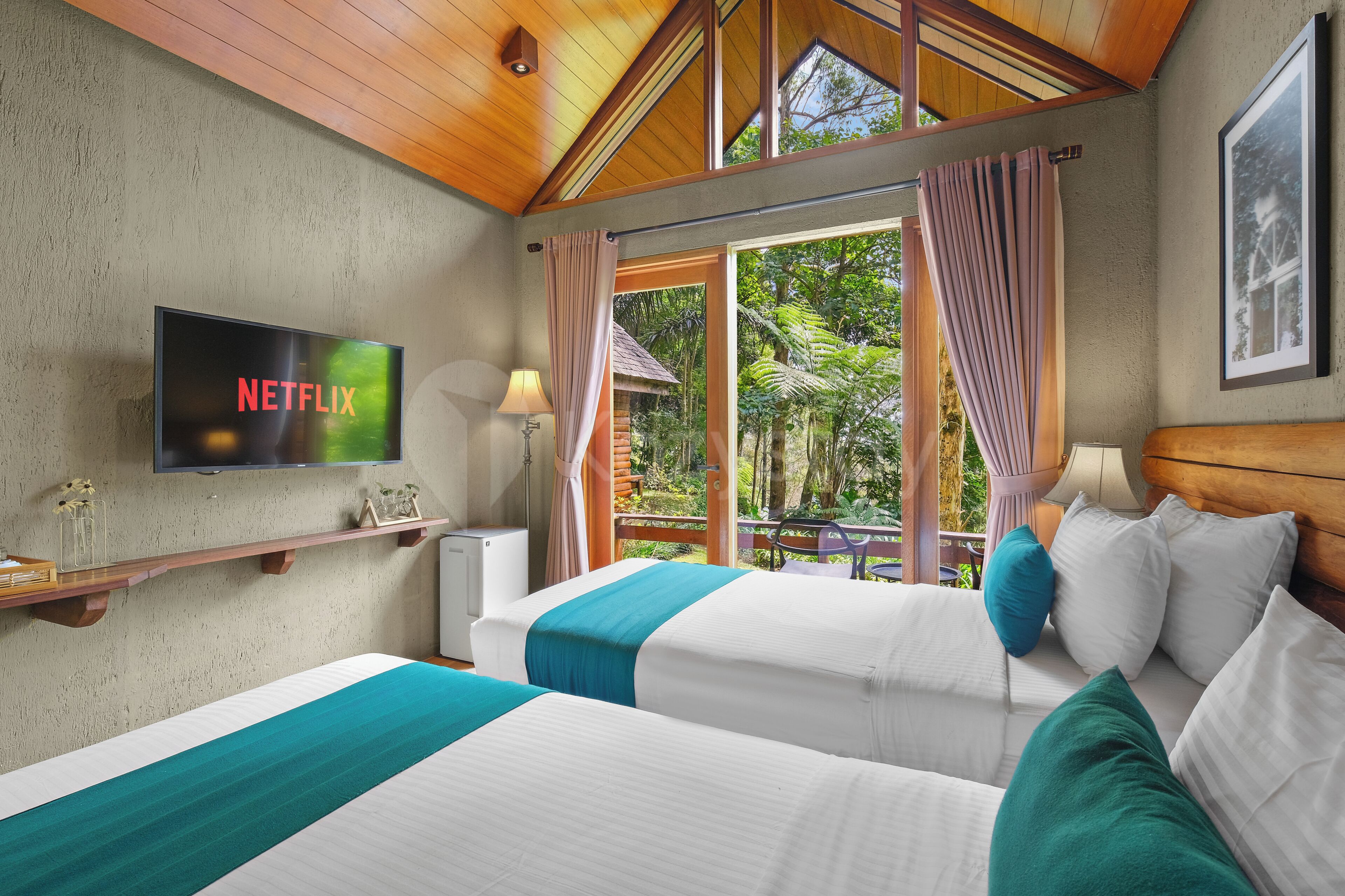Ein Hotelzimmer mit zwei Betten, einem Fernseher, einem Balkon mit Vorhängen und Blick auf Grünflächen.