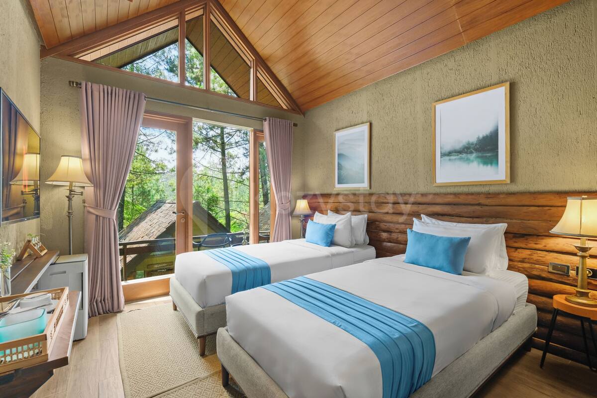 A room with two beds, a wooden ceiling, a large window with curtains, a framed picture on the wall, and a small table with a lamp.