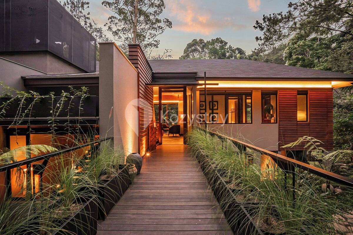 A modern house with a wooden deck leading to a glass door entrance, surrounded by lush greenery.