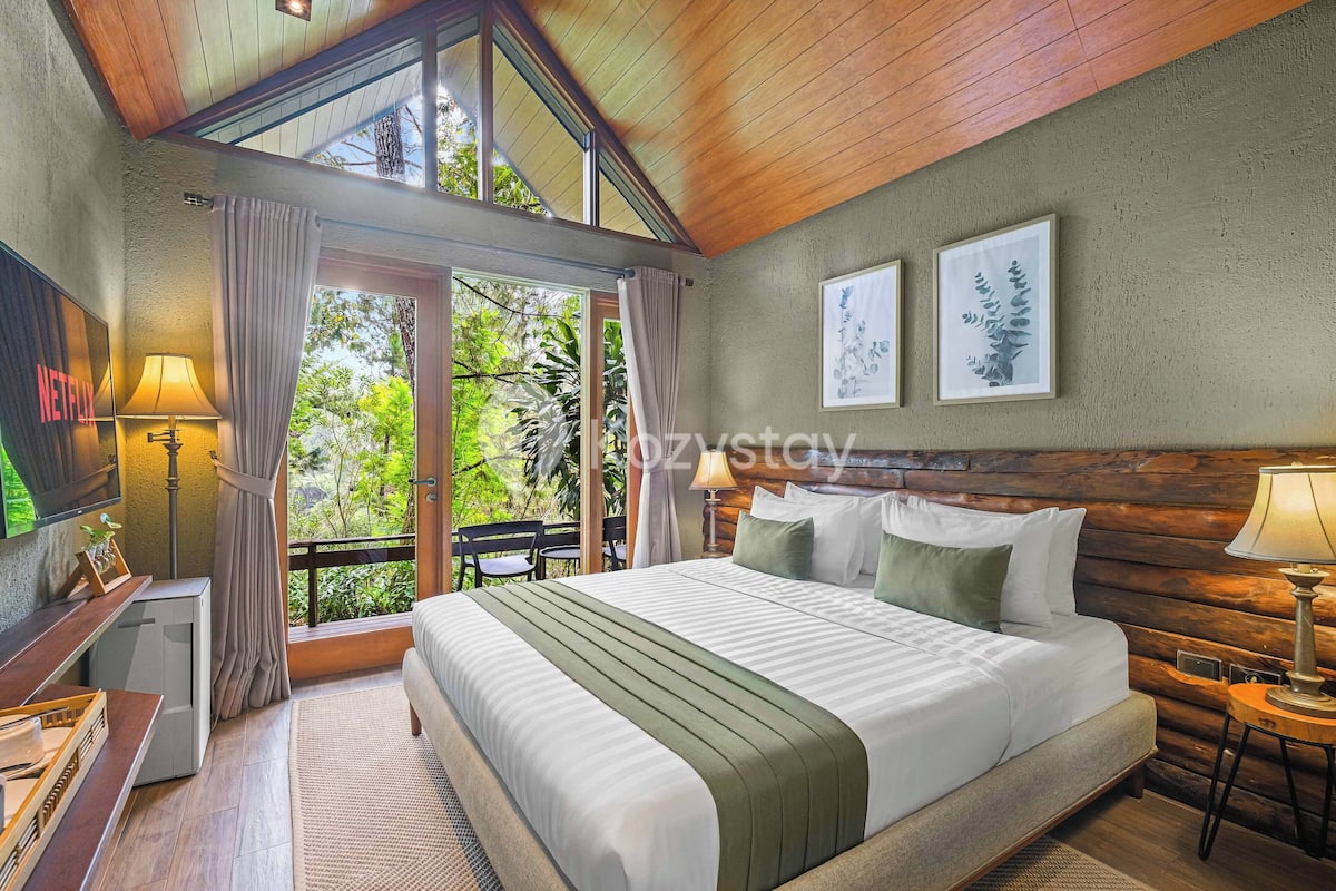A hotel room with a large bed, a TV, a desk, and a balcony with a view of greenery.