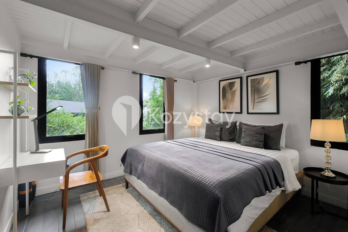 A modern bedroom with a large bed, a desk, and a chair. There are two windows with curtains, a ceiling with exposed beams, and a small table with a lamp.