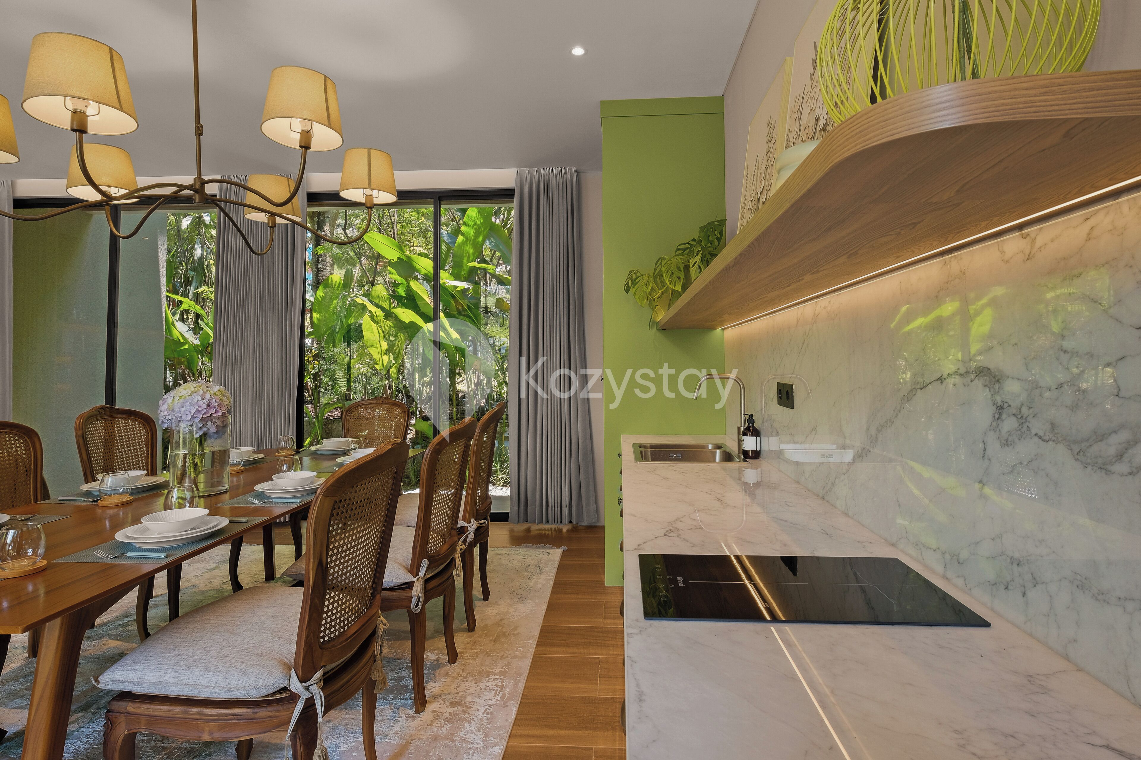 A modern dining area with a wooden table set for six, a chandelier, and a view of greenery through large windows.
