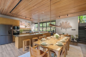 A modern kitchen with wooden cabinetry, a central island, and a dining area set for six. - Taman Wisata Bougenville by Kozystay (Banjaran)