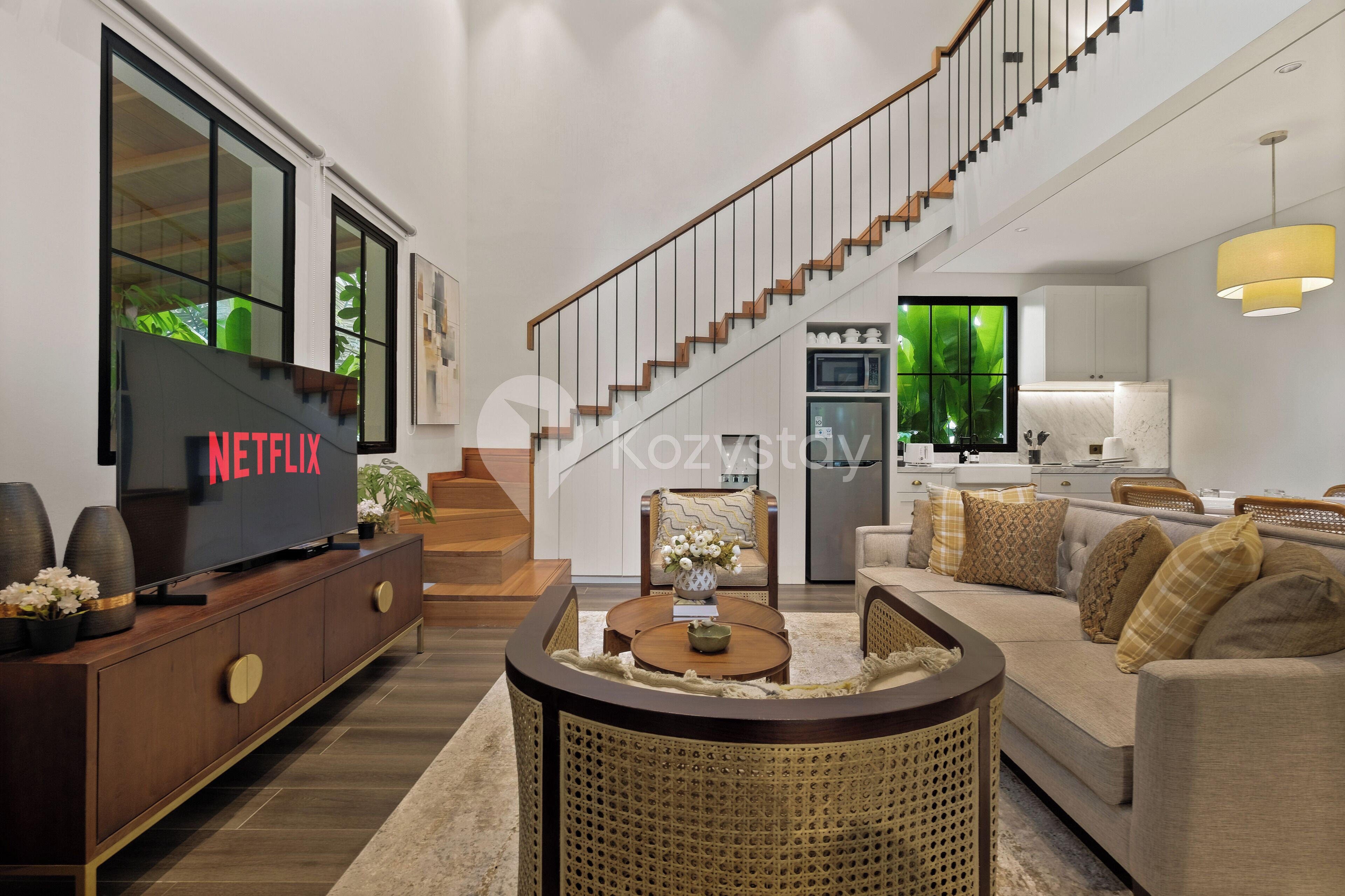A modern living room with a large flat-screen TV, a staircase, and a kitchen area.