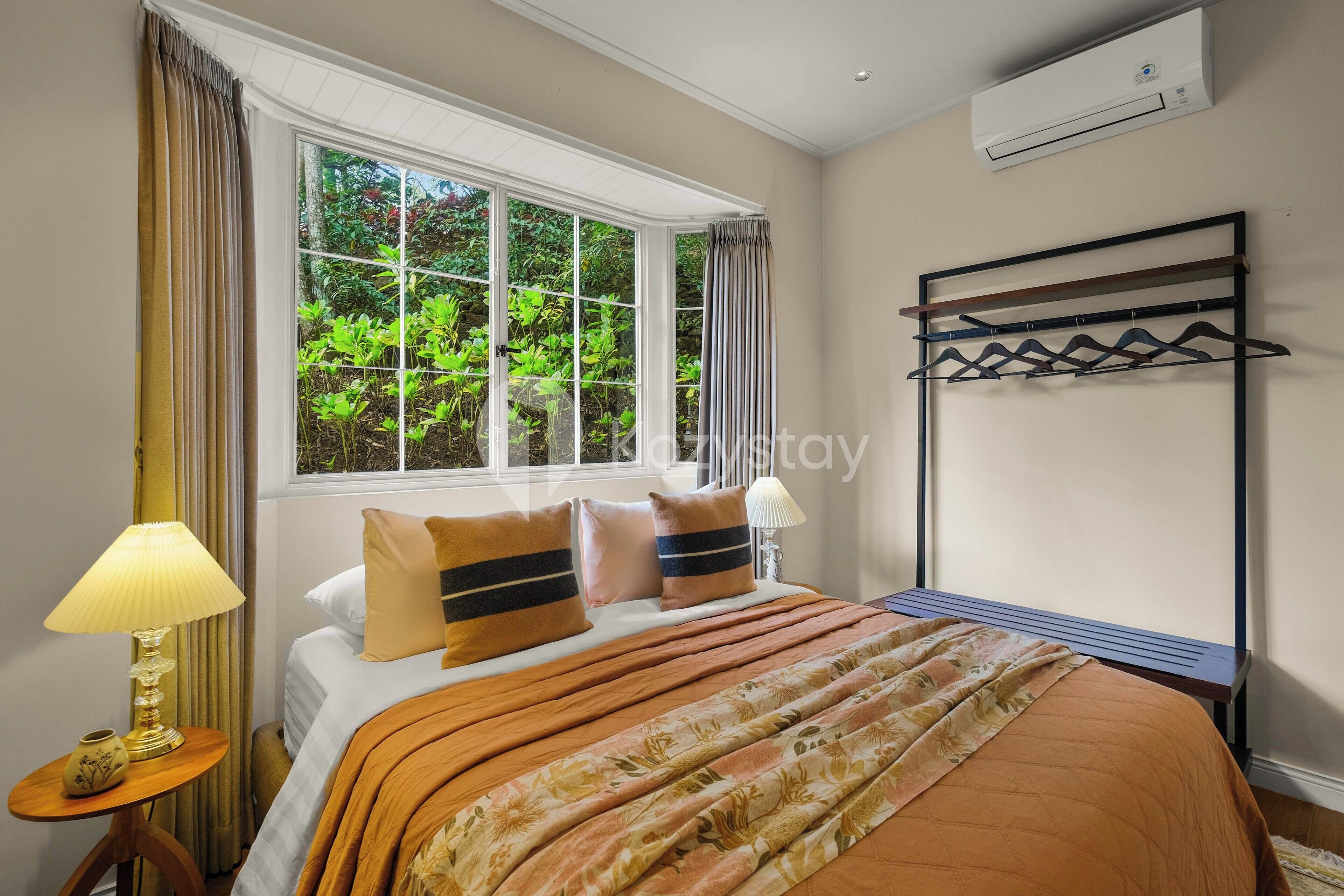 A hotel room with a large window, a bed with a floral bedspread, a bedside table with a lamp, and a wall-mounted drying rack.