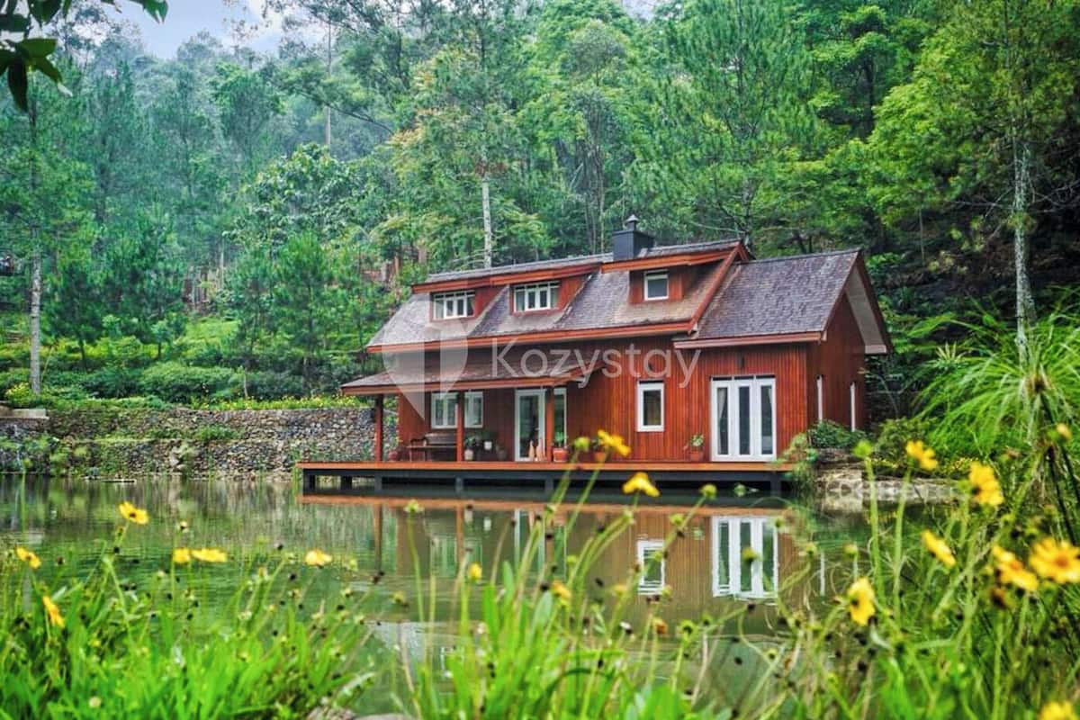 A wooden house with a balcony, situated by a lake surrounded by lush greenery.