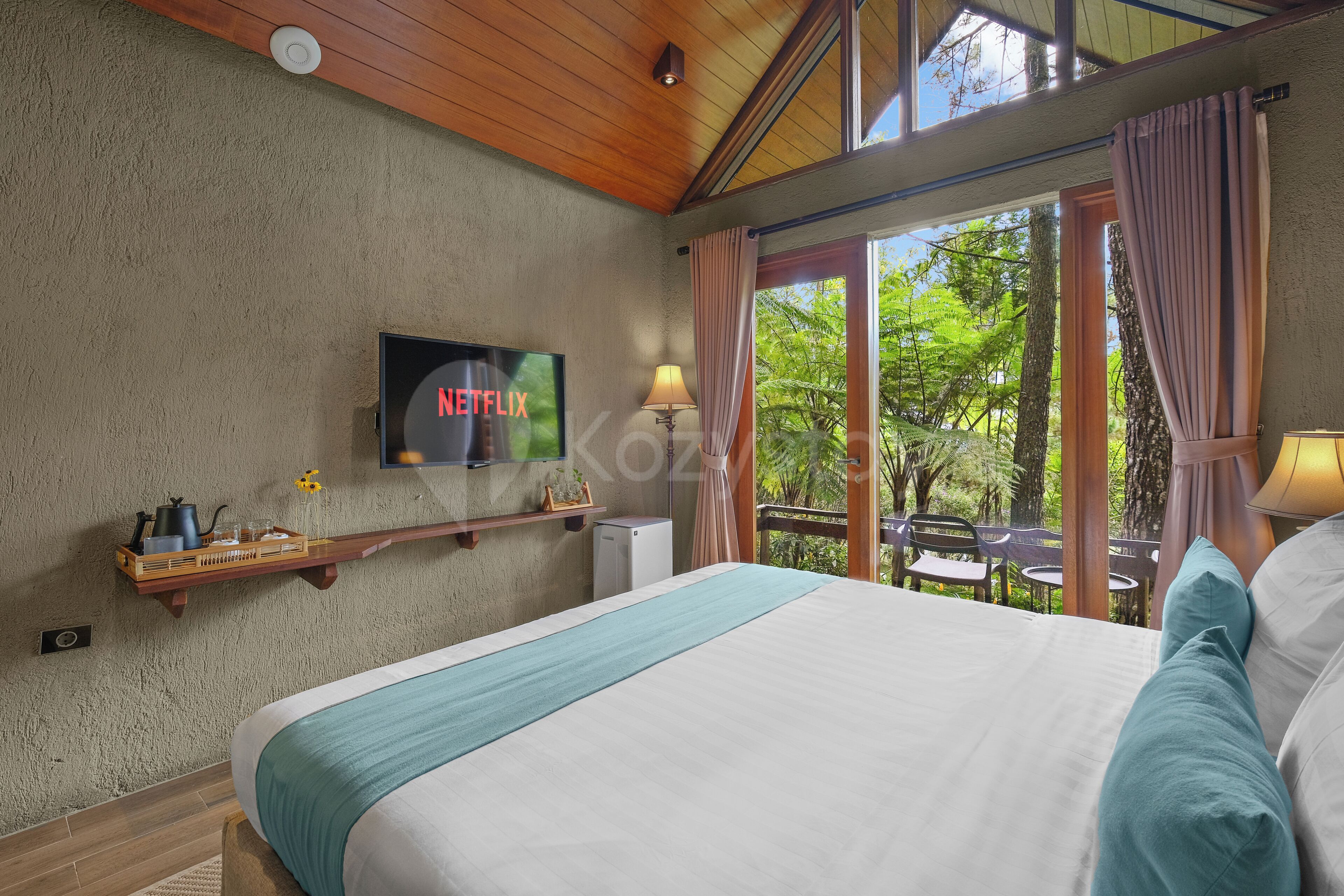 A hotel room with a bed, a TV mounted on the wall, a small table with a lamp, and a view of the outdoors through large windows.