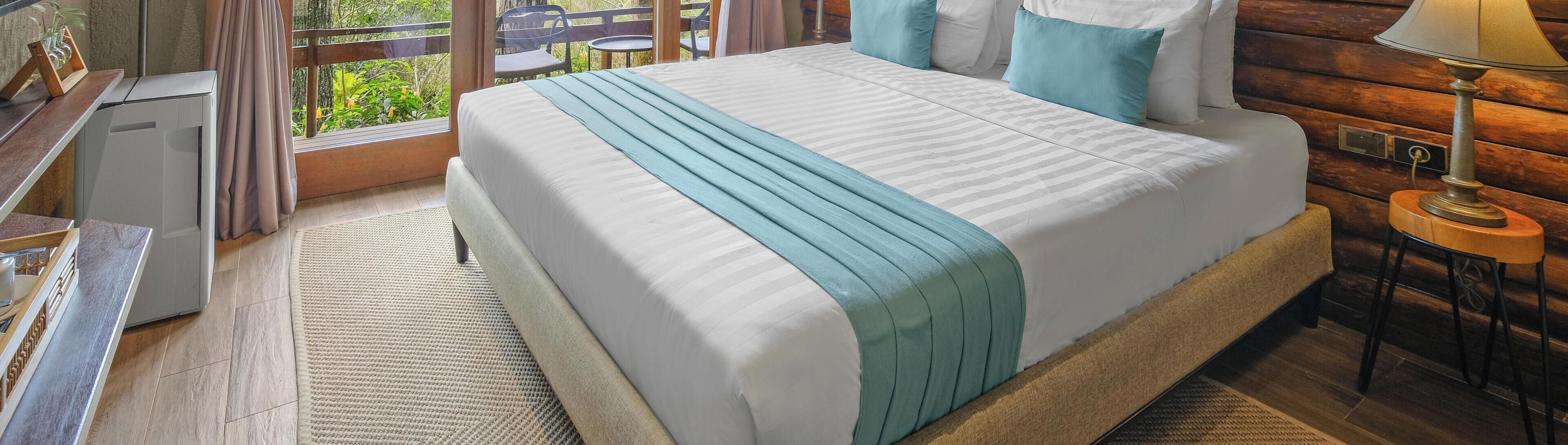 A neatly made bed with a striped blanket, a wooden nightstand with a lamp, and a view of greenery through a window.