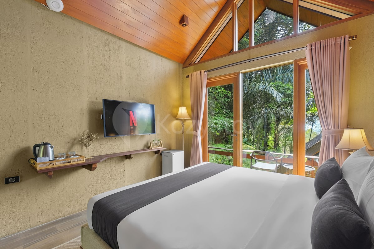 A bedroom with a large bed, a television, a small table with a lamp, and a view of greenery through the window.